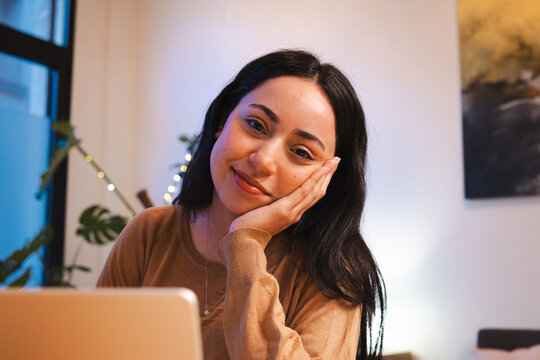 A young woman with dark hair rests her chin on her hand, smiling gently while sitting at a desk with a laptop.