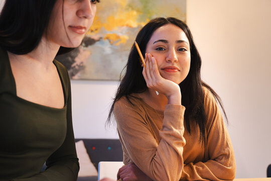 Two young women are studying together, one holding a pencil and resting her chin on her hand, the other looking thoughtfully into the distance.