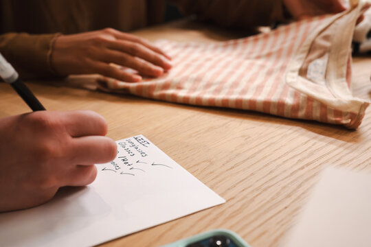 A person's hands are shown as one writes a list on paper while the other smooths a striped garment, suggesting preparation or organization.
