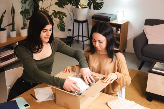 Two young women are unpacking a cardboard box filled with items, reviewing a handwritten list on the table.