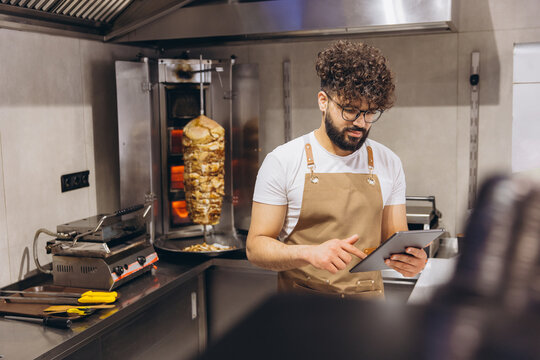 Arab man chef managing online orders on tablet in fast food restaurant kitchen with shawarma cooking
