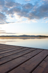 Fototapeta premium Vertical landscape of a serene lake at sunrise, viewed from a wooden deck in the foreground. Calm water reflects soft morning light, with mountains rising in the middle distance and colorful sky above
