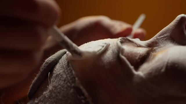 Closeup cinematic sequence of a relaxing spa facial treatment with foaming mask applied by esthetician brushes on peaceful client skin under warm ambient lighting