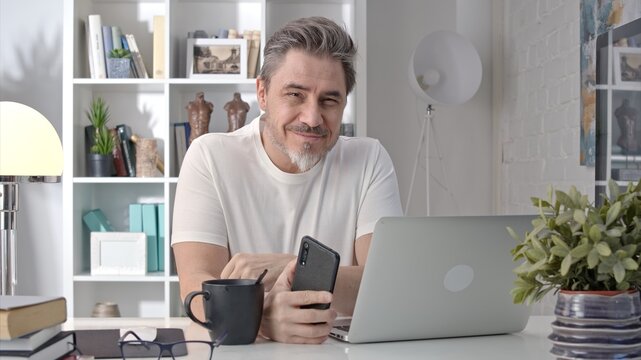 Older white man in glasses sitting at desk using phone. Home office, businessman working from home, morning coffee, checking social media news feed.