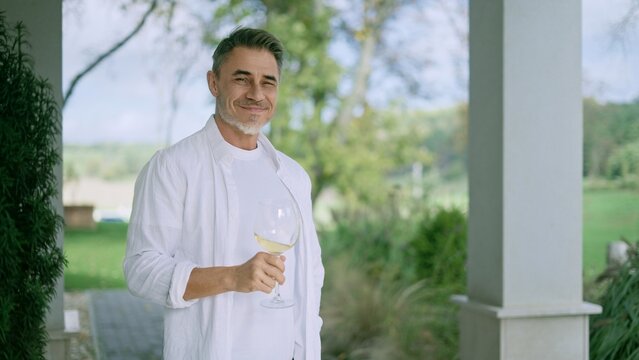 Portrait of mature man relaxing on country house terrace, holding glass of white wine. Satisfied landowner enjoying peaceful view of ranch on porch of farmhouse home. Smiling mid adult male in white.