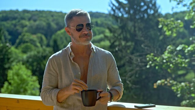 Businessman starting the day with morning coffee and checking phone. Portrait of middle aged man in eyeglasses and casual cloth outdoor on home balcony. Spring or summer. Happy satisfied smile.