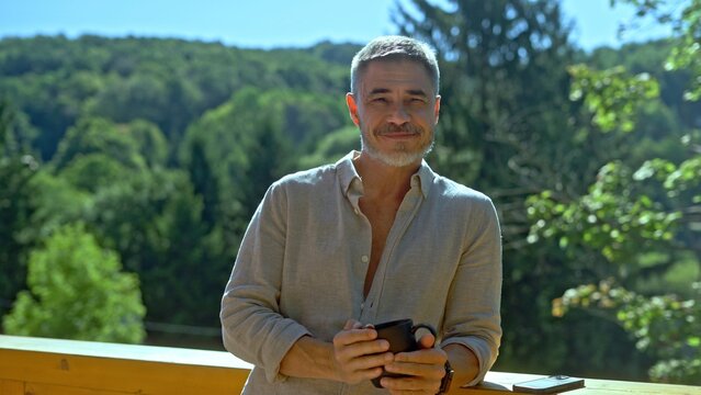 Portrait of middle aged man with gray hair in casual cloth outdoor on home balcony. Mid adult male starting the day with morning coffee. Spring or summer. Happy satisfied smile.
