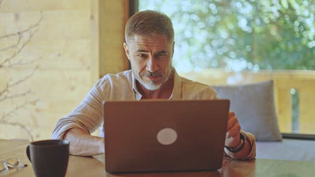 Businessman remote working on laptop online in home office, concentrating, thinking. Middle aged man sitting at desk in living room. Spring or summer. Entrepreneur on getaway in wooden cabin.