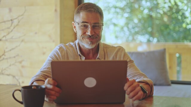 Happy businessman remote working on laptop online in home office, smiling. Middle aged man in eyeglasses sitting at desk in living room. Spring or summer. Entrepreneur on getaway in wooden cabin.