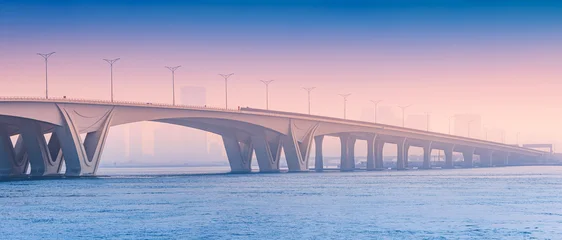 Fototapeten Abu Dhabi bridge providing a modern architectural link over calm water during an atmospheric morning in Abu Dhabi. Urban silhouettes visible in the distance  © EdNurg
