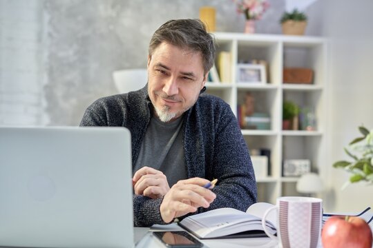 Bearded man working online with laptop computer at home sitting at desk. Home office, browsing internet, study room. Portrait of mature age, middle age, mid adult man in 50s.