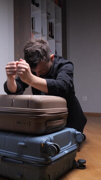 Vertical video man struggling with a stacked pile of suitcases, looking stressed and frustrated while preparing for a trip, experiencing packing difficulty and travel anxiety at home