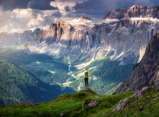 Naklejka premium Sporty woman standing on a grassy hilltop with stunning view of the alpine mountains at sunset in summer. Scenery with girl, dramatic clouds over rocks and green valleys. Hiking in Dololmites, Italy