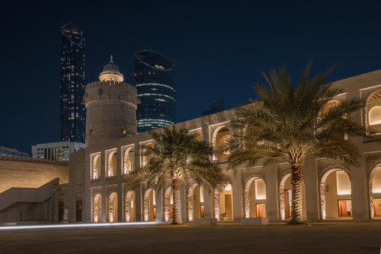 The historic Qasr Al Hosn fort illuminated at night with palm trees and arched arcade, contrasting with modern glass skyscrapers in the background, Abu Dhabi, United Arab Emirates 