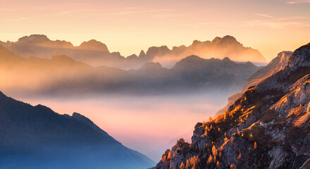 Naklejka premium Mountains in low clouds at beautiful sunset in autumn in Dolomites, Italy. Landscape with alpine mountain hills in fog, orange trees and grass in fall, colorful sky with golden sunbeams. Aerial view