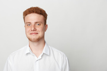 Obraz premium Young man with red hair and beard wearing white shirt smiles gently against plain light background. Portrait of confident male with casual style and natural expression.
