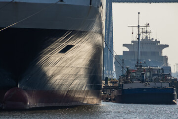 Naklejka premium A massive cargo ship bow dominates the frame while a supply boat maneuvers beside it in a busy industrial Klaipeda port, conveying maritime transport, heavy industry and harbor operations up close. 