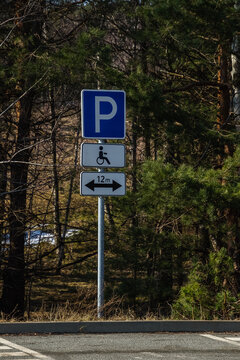 Handicap and parking signs mounted on a metal pole beside a forested roadside, showing a blue P, wheelchair symbol and 12m directional arrow to indicate accessible reserved parking spaces nearby.
