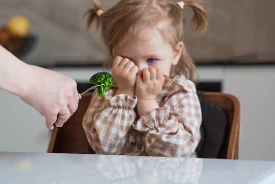 Mom is spoon-feeding a little girl broccoli. The child refuses to eat. Baby food. Healthy food.