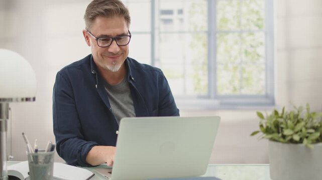 Casual businessman working with laptop computer in home office, happy, smiling. Portrait of middle age man in 50s, white caucasian, gray hair.