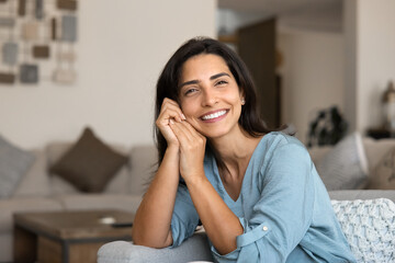 Calm Latina woman sitting comfortable on cozy sofa, looks at camera with contented smile, enjoying...