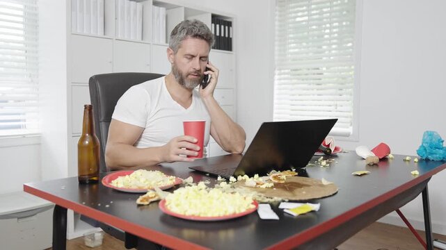 Man using laptop in stressful work situation. Office worker distracted by food while working. Man typing on laptop at cluttered desk. Office worker working with unhealthy lifestyle.