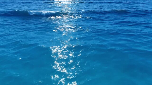 Aerial drone perspective above ocean waves vibrant blue gradient water luminous sunlight reflections shimmering across surface soft white foam accents natural wave rhythm smooth cinematic camera glide