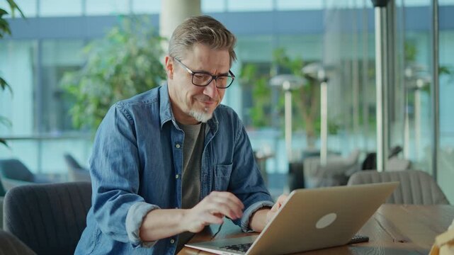 Happy middle age businessman using laptop computer in office. Older casual man working online on terrace of cafe, smiling.
