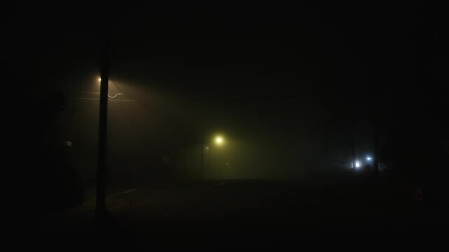 Empty urban street at night illuminated by sparse street lights whose glow diffuses through dense fog, creating a tense and cinematic atmosphere of silence and isolation