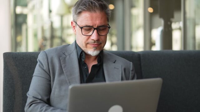 Businessman sitting on couch in hotel, office lobby, working with laptop computer. Mature age, middle age, mid adult man in business casual, confident happy smiling.