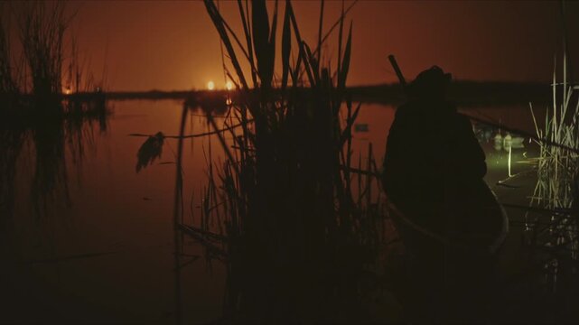 Silhouette of a hunter in a boat searching for prey in a swamp during the dark orange night