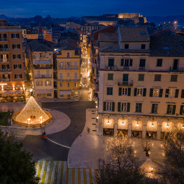 op-down aerial view of a busy intersection in Corfu Old Town with the Liston, Christmas tree, and Old Fortress in the background during a festive blue hour evening