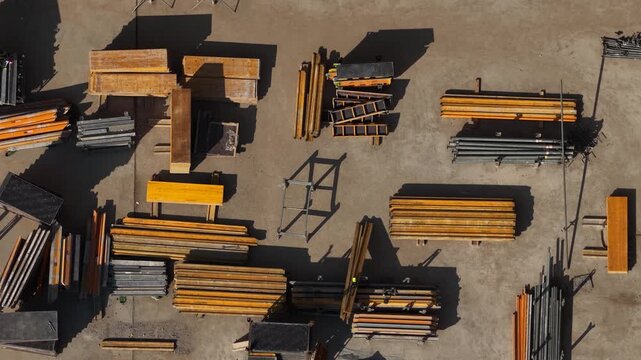 Drone shot of neatly arranged construction materials including wooden formwork panels, steel pipes, and equipment on a clean concrete surface at a building site.