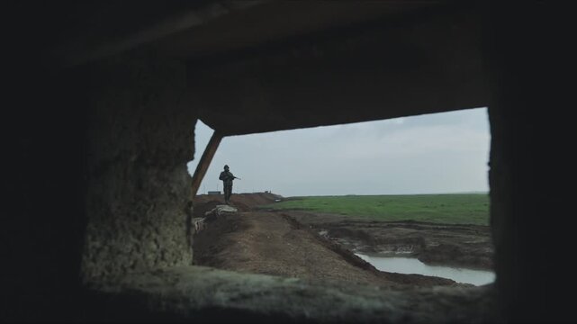 Soldier with a rifle patrolling a muddy trench seen from inside a concrete bunker during wartime
