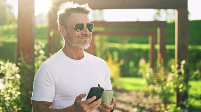 Happy mature man using smartphone drinking coffee in garden during beautiful sunrise. Portrait of senior male in sunglasses enjoying morning moment of relaxation on home terrace. , smiling .