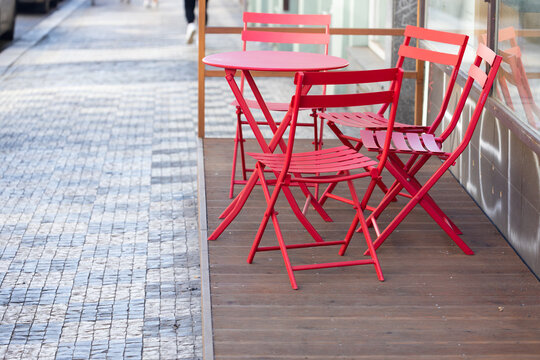 Red folding chairs and round table on wooden deck outside a cafe, with cobblestone pavement and blurred pedestrians in the background