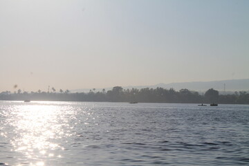Naklejka premium Beautiful morning landscape of a wide tropical river with sunlight reflecting on the water surface and silhouettes of traditional boats near a lush palm tree coastline under a clear misty sky.