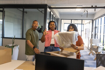 Naklejka premium African American coworkers unpacking bubble-wrapped monitor at open-plan office desk with boxes