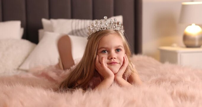 Beautiful little girl with crown dreaming about becoming queen on bed at home
