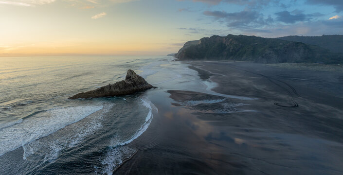 Aerial view of waves crashing against a rock formation on the beach at Karekare Beach in Auckland, New Zealand. The tide is low, and the sand is black.