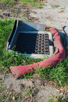 A metal storm drain grate surrounded by a red silt sock on sandy soil and grass, illustrating erosion control, stormwater protection and roadside drainage maintenance at a construction or urban site.
