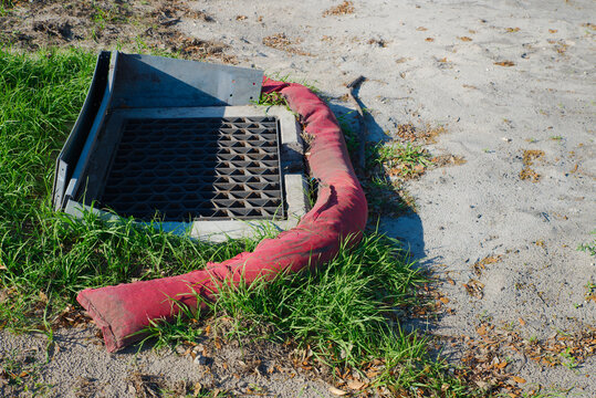 A metal storm drain grate surrounded by a red silt sock on sandy soil and grass, illustrating erosion control, stormwater protection and roadside drainage maintenance at a construction or urban site.
