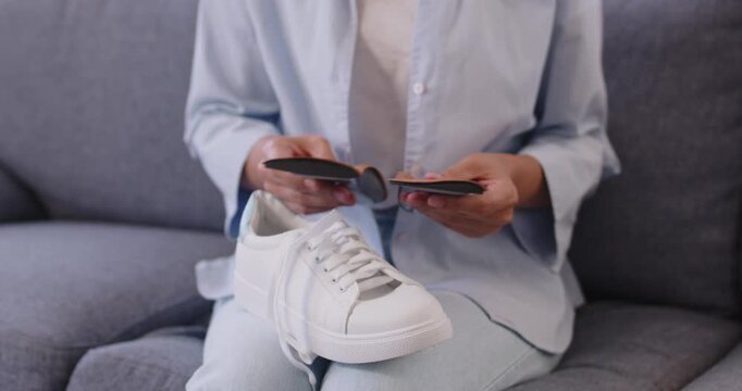 Woman examining orthopedic insoles for sneakers to improve foot support and flatfoot correction. Person sitting on sofa at home to compare individual inserts beside white sport shoes, check arch shape