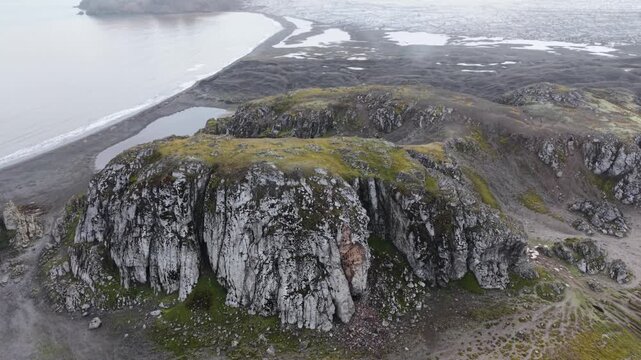 Aerial circles a prominent moss-covered basaltic rock at Elephant Point, Livingston Island, revealing the black sand beach, lagoon, and rugged Antarctic landscape