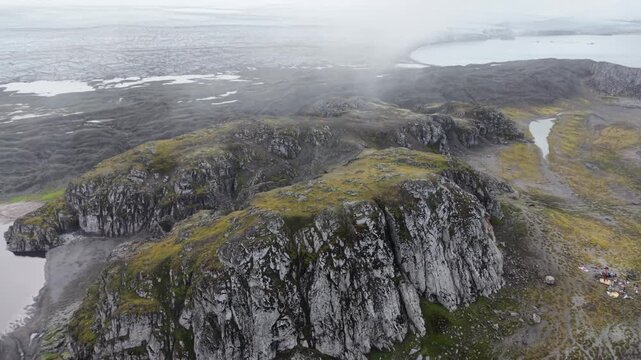 Aerial camera circles a prominent moss-covered basaltic rock outcrop at Elephant Point, Livingston Island, Antarctica revealing rugged Antarctic landscape and distant southern elephant seal colonies