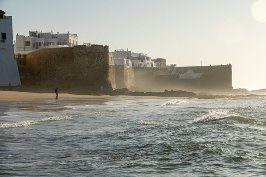 Silhouette of a man fishing on the beach, with the walls of the medina in the background, in Asilah, Morocco
