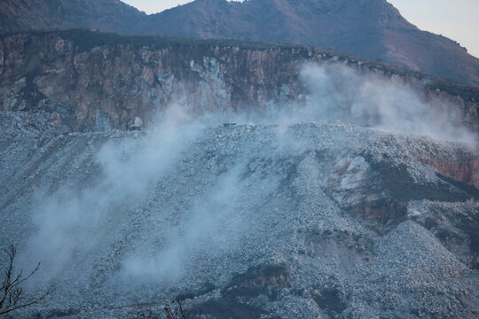 A wide-angle view of a white dust plume rising from a marble excavation site in the mountainous terrain of Buner, Pakistan, framed by winter trees.