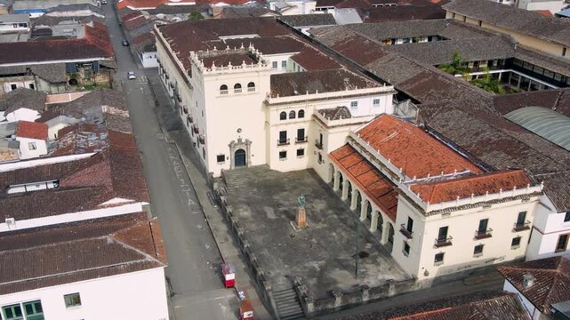 Drone aerial of Popay&aacute;n showing a pale facade building, a raised stone plaza with central statue, red clay tile roofs and a nearly empty street.