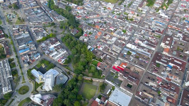 Drone pans over Popayan showing a tree-lined park, grid of red-tiled rooftops, white buildings and a circular roundabout from an overhead perspective.