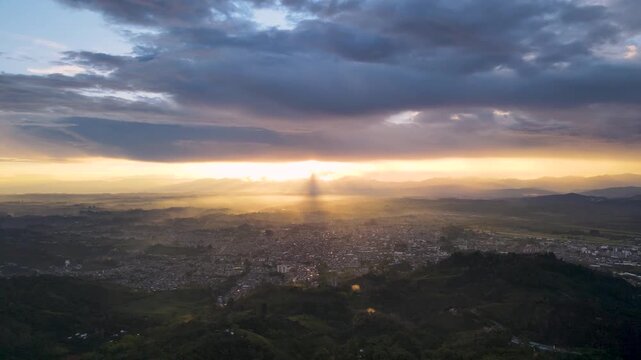 Aerial view of Popayan with golden sunbeams piercing low clouds, mist over green hills and the compact city grid spreading toward the horizon.